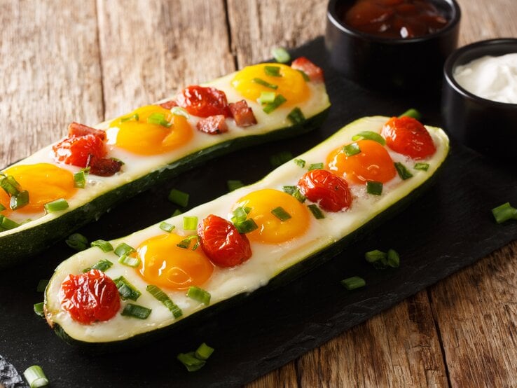 Zwei Spiegelei-Zucchini-Boote mit Kirschtomaten, eins mit Speck und beide mit Frühlingszwiebeln garniert auf einem schwarzen Brett neben zwei Schüsseln mit Soßen, vor einem Holzhintergrund. 