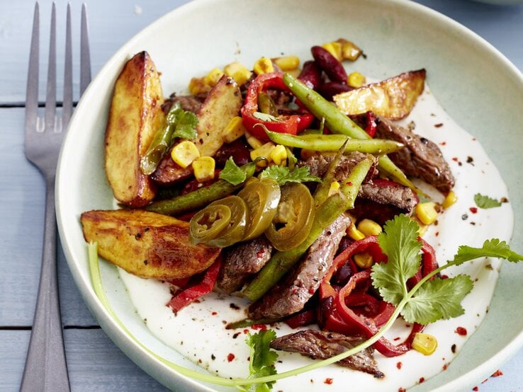 Mexikanisches Geschnetzeltes mit angerösteten Bohnen, Kartoffeln, Mais, Chili und frischem Koriander, serviert auf einem weißen Teller auf hellblauem Holztisch. 