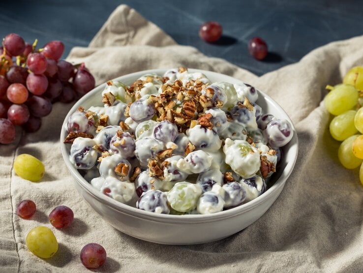Auf einem Tuch steht eine runde Schüssel. In der Schüssel ist ein Weintrauben-Salat mit Nüssen. Drumherum liegen dunkle und helle Weintrauben.