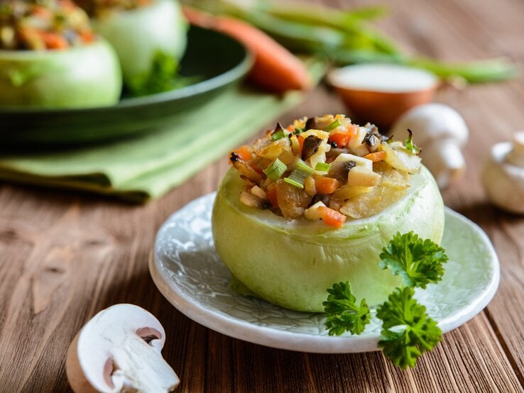 Frontalansicht: eine gefüllte Kohlrabi mit Champignons und Gemüse. Im Hintergrund sind zwei weitere Exemplare zu sehen. Drumherum liege drei frische Champignons.