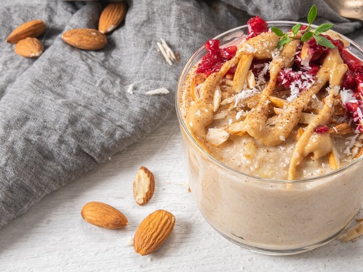 Ein Glas mit dem Mandelporridge, Himbeeren und Kokosraspeln auf einer hellen Tischplatte mit einem grauen Tuch drum herum.