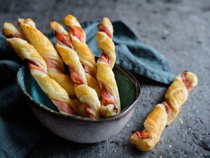 Blätterteigstangen mit Speck un einer Schüssel auf dunklem Untergrund. 