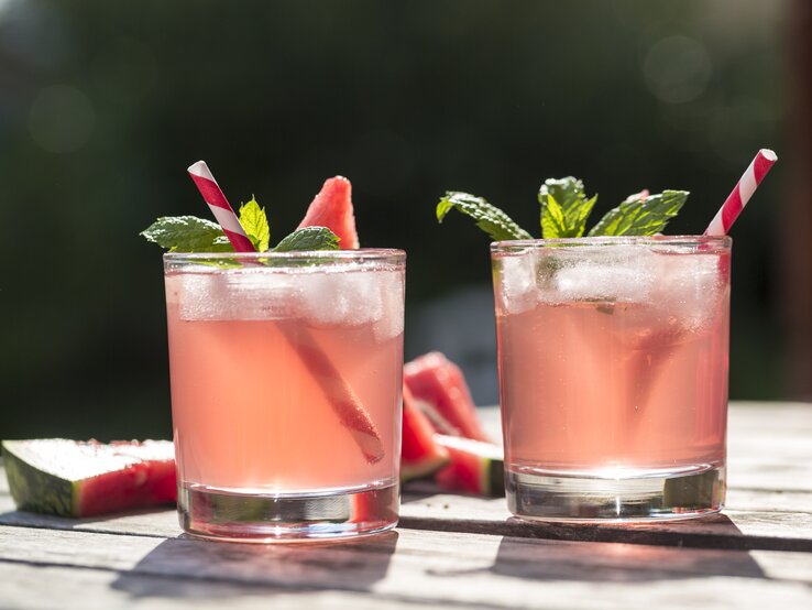 Zwei Gläser gefüllt mit rosafarbenem Wasser-Melonen-Hugo – Dekoriert mit Frucht, Minze und Strohalmen.  | © Getty Images/Westend61