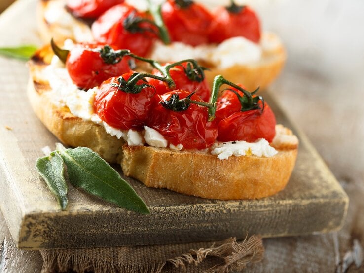 Gegrillte Bruschetta mit geschmolzenen Tomaten und Salbei auf hellem Holzbrett und grauer Stoffserviette. Im Hintergrund unscharf eine weitere. Frontalansicht.