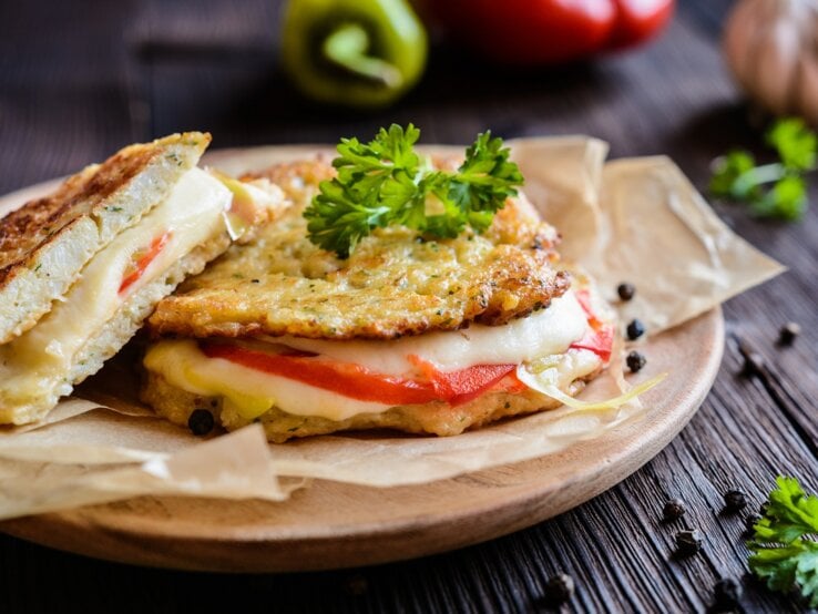 Die Low-Carb-Panini mit Mozzarella und Tomate auf einem Brett. Dahinter verschiedene Zutaten auf einem hölzernen Untergrund.
