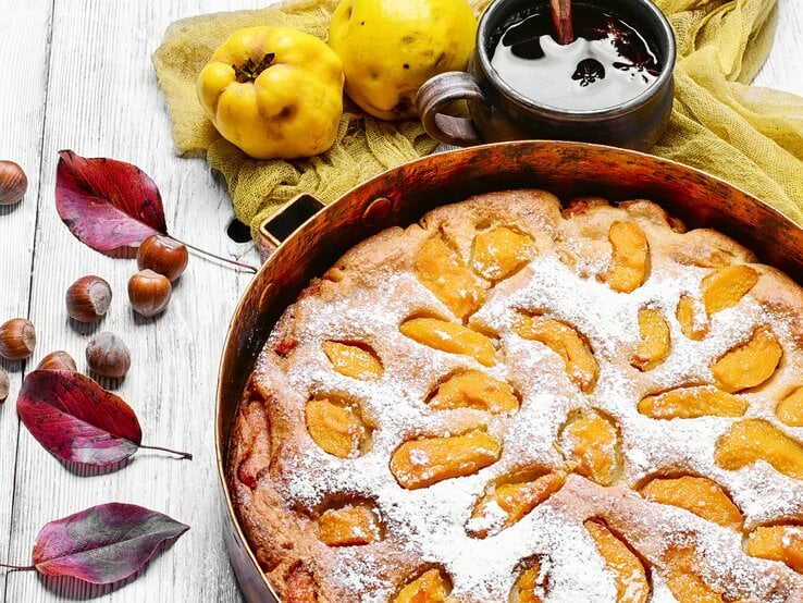 Draufsicht: ein runder Kuchen mit Quitten, der noch in der Backform ist. Er liegt auf einem hellen Untergrund aus Holz. Daneben liegen Blätter von einem Baum, Haselnüsse und Quitten. Neben dem Kuchen steht eine dunkle tasse mit einer dunklen Flüssigkeit.
