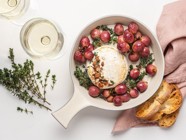 Eine feuerfeste Form mit einem Ofenkäse mit Weintrauben, Thymian, Honig und Nüssen, daneben stehen zwei Gläser mit Weißwein und einer altrosa Stoff-Serviette zur Dekoration.