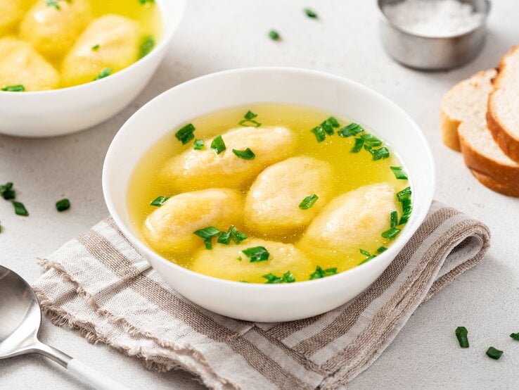 Draufsicht, schräg: Zwei weiße Schüsseln mit einer bayerischen Grießnockerlsuppe stehen auf einem hellen Untergrund. Daneben liegen ein Löffel, zwei Scheiben Weißbrot und eine Schale mit Salz.