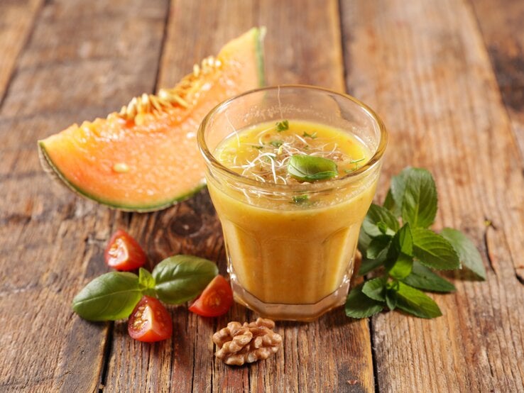 Ein Glas Honigmelonen-Gazpacho mit Sprossen und Basilikum bestreut, daneben liegen ein Stück Melone, Tomaten und Walnüsse.