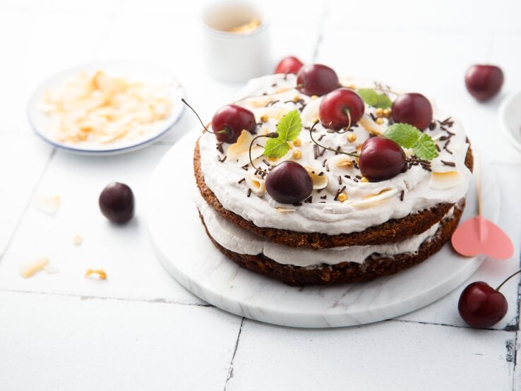 Schüttelkuchen nach Schwarzwälder-Kirsch-Art auf weißem, hölzernem Untergrund – umgeben von einigen Zutaten. 