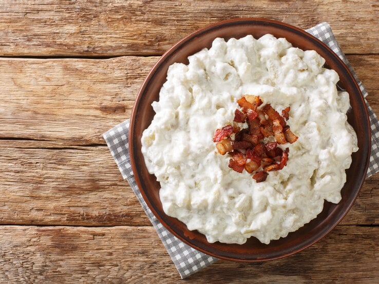 Auf einem Untergrund aus Holz liegt ein grau-weißes kariertes Tuch. Auf dem Tuch steht eine Schale mit Spätzle mit Feta und Speck. Es handelt sich um ein Gericht aud der Slowakei.