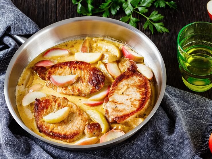 Ein silberner tiefer Teller mit drei Schweinemedaillons und der Apfelweinsoße mit den Apfelspalten. Um den Teller herum frischer Apfel, Petersilie, ein dunkelgraues Tuch und ein paar grüne Gläser.