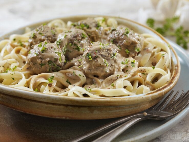 Ein tiefer Teller mit Köttbullar und Fettuccine in einer hellen Soße, bestreut mit gehackter Petersilie.