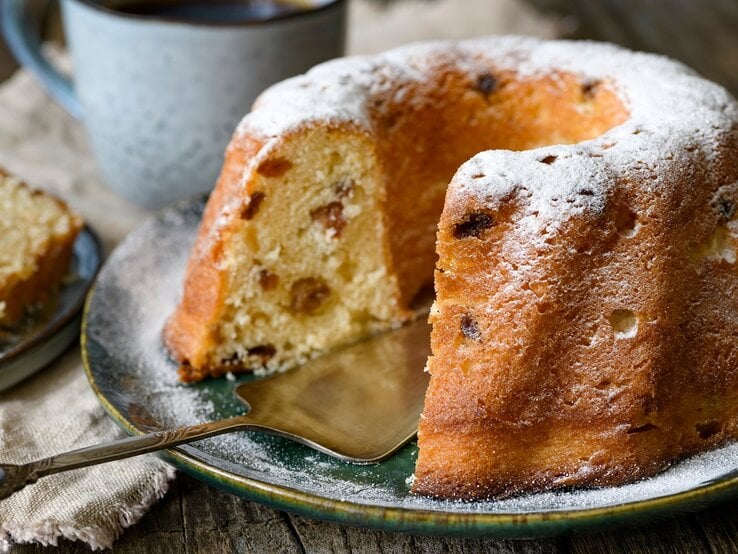 Frontalansicht: Ein angeschnittener Hefe-Gugelhupf steht auf einem Teller. Dahinter stehe eine Tasse und ein Stückchen Kuchen.