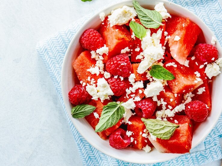 Wassermelonenslata mit Minze, Himbeeren und Feta in einer weißen Schale auf einem blauen Tuch. Auf einem hellen Tisch mit zwei Holzgabeln an der rechten Seite.