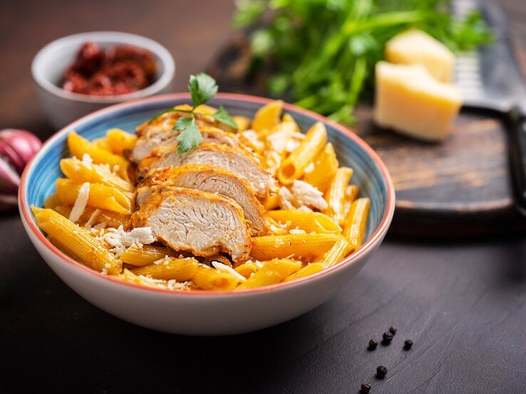 Cajun One-Pot-Pasta mit Hähnchen in einem bunten tiefen Teller. Dahinter eine Schale mit Tomaten, Gewürze und Parmesan. 