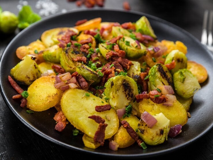 Ein schwarzer Teller mit Gröstl mit Rosenkohl auf einem schwarzen Tisch.