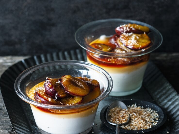 Zwei Portionen Kokospudding mit karamellisierten Zwetschgen stehen auf einem gemusterten schwarzen Teller auf einem Holztisch. Der Pudding ist angerichtet mit Kokosflocken.