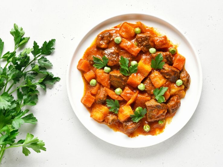 Auf einem weißen Untergrund steht ein weißer Teller. Auf diesem ist ein zartes Lammragout mit Kartoffeln. Daneben liegen einige Zweige Petersilie.