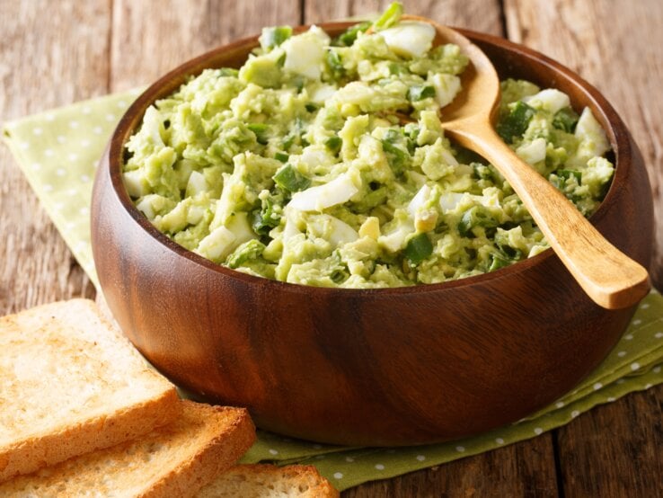 Avocado-Eiersalat mit Lauch und Joghurt in Holzschüssel auf grüner Serviette und Holztisch. Oben auf Holzlöffel. Links am Bildrand geröstetes Toastbrot. Draufsicht.