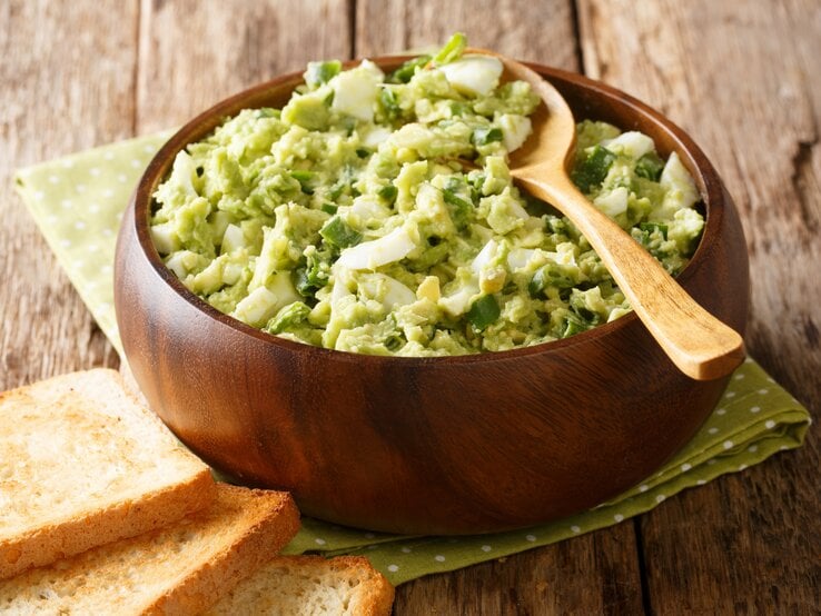 Avocado-Eiersalat mit Lauch und Joghurt in Holzschüssel auf grüner Serviette und Holztisch. Oben auf Holzlöffel. Links am Bildrand geröstetes Toastbrot. Draufsicht. | © Shutterstock / AS Food studio