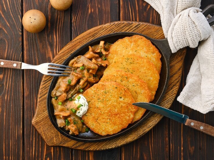 Eine gusseiserne Pfanne mit den Haferpuffern an einem Pfifferlingsrahm von oben fotografiert auf einem dunklen, rustikalen Holztisch. Außerdem sind auch Messer, Gabel und ein helles Tuch mit auf dem Bild.  