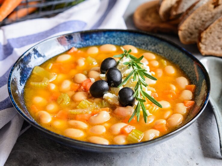 Bohnensuppe in einer blauen Schale auf grauem Tisch. Im Hintergrund ein Brotkorb, ein blau-weißes Tuch und ein Bund Karotten.