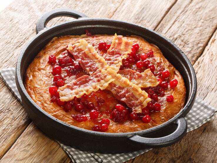 Eine Pfanne mit dem Äggakaka, dem schwedischen Eierkuchen garniert mit kross gebratenen Bacon-Scheiben und Preiselbeeren, auf einem karierten Untersetzer vor einem Holzhintergrund. 