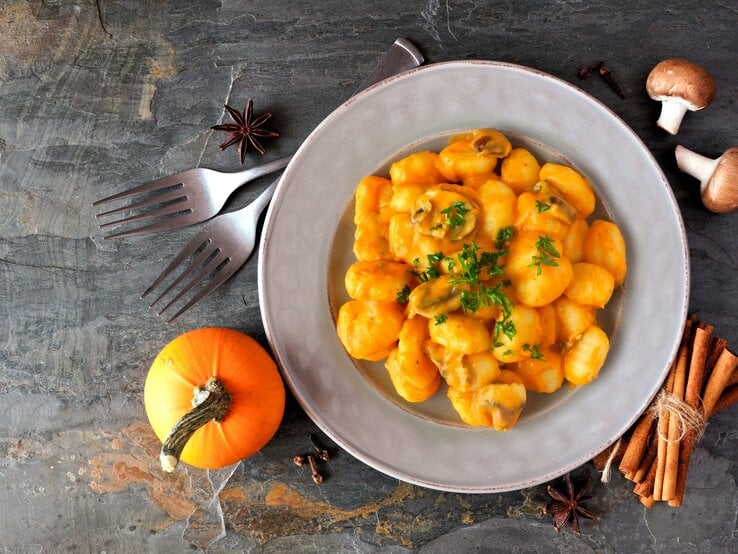 Eine Portion Kürbis-Gnocchi mit Kürbis-Pilz-Soße in einem weißen Teller steht auf einem Untergrund aus Stein. Um den Teller herum liegen Zimt, Sternanis, ein Kürbis, Pilze und zwei Gabeln. | © Getty Images / jenifoto