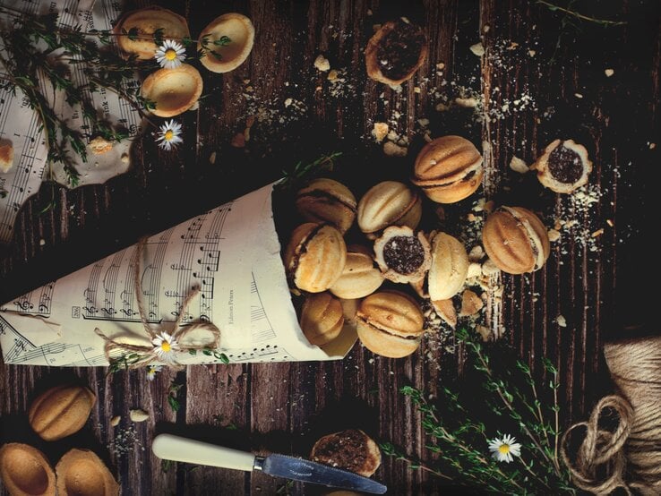 Eine Papiertüte aus Notenblättern gefüllt mit vielen Stücken der polnischen Zaubernüsse Orzeszki, einige im ganzen Stück, andere sind geöffnet, sodass man die Schokoladenfüllung sehen kann. daneben sind weitere Notenblätter, Blumen und ein Messer zu sehen, vor einem Holzhintergrund von oben.  | © StockFood / Wrobel-Sterianos, Anna