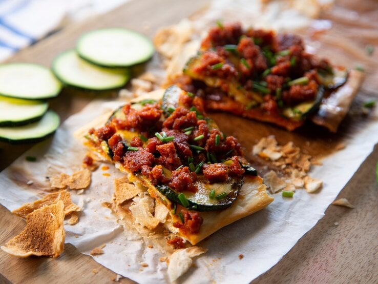 Spanische Maismehlpizza Coca Mallorcina auf Backpapier und Holzbrett. Daneben Zucchinischeiben. Draufsicht.