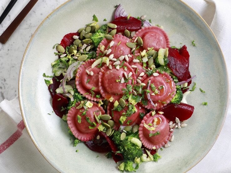 Salat mit Rote-Bete-Tortellini, Zwiebeln, frischer Rote Bete, Brokkoli, Kernen und Kräutern, serviert in einer hellen Schale auf einem Marmortisch, von oben fotografiert. 