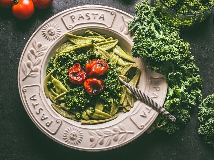 Ein Teller Grünkohl-Pasta mit Zitrone und Tomaten in der Draufsicht, daneben frischer Grünkohl und ganze Tomaten.