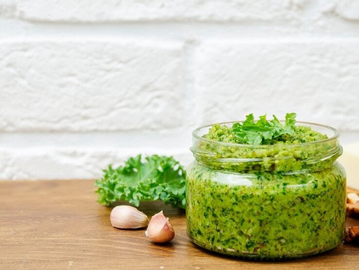 Ein Glas Grünkohlpesto, daneben Knoblauchzehen, Walnüsse und frischer Grünkohl. | © Adobe Stock/ nata_vkusidey 