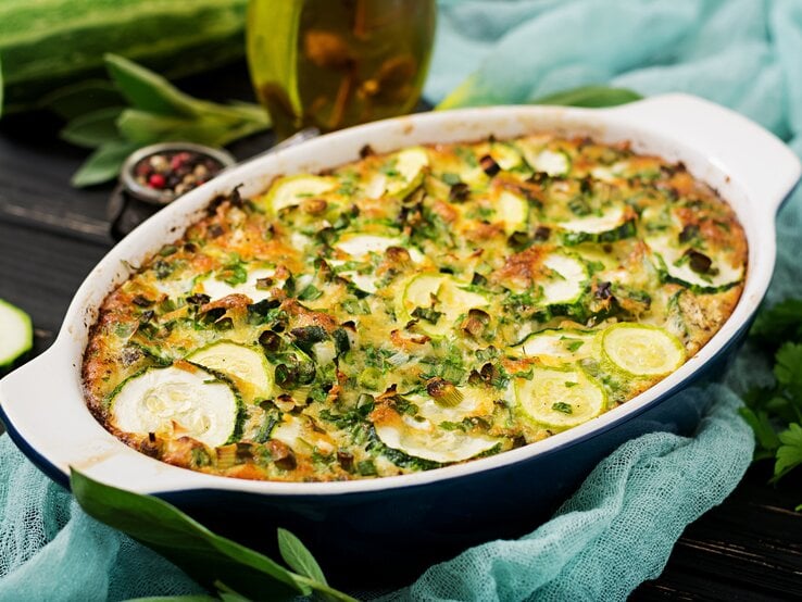 Kartoffel-Lauch-Gratin mit Zucchini in dunkelblauer Auflaufform, angerichtet auf einem blauen Tuch auf dunklem Holz. Umgeben ist die Speise von den Zutaten. 