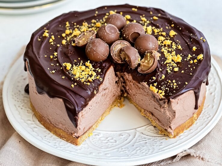 Ein weißer Teller mit einem angeschnittenen Baileys-Käsekuchen mit Schokoglasur auf einem braunen Tischtuch vor einem hellen Hintergrund.  | © StockFood / Parissi, Lucy