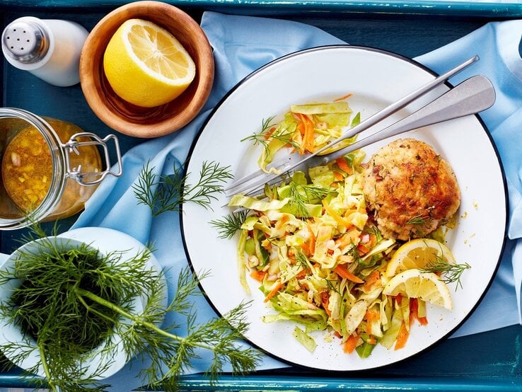 Forellen-Frikadell mit Spitzkohlsalat und Dillgarnitur und weißem Teller mit schwarzem Rand auf blauem Tuch, blauem Tableet und Holztisch aus der Vogelperspektive. Außerdem Messer und Gabel auf dem Teller. Links halbe Zitrone in Tonschale, daneben weißer Salzstreuer, Einmachglas mit Brühe und weißes Kännchen mit Dill, 