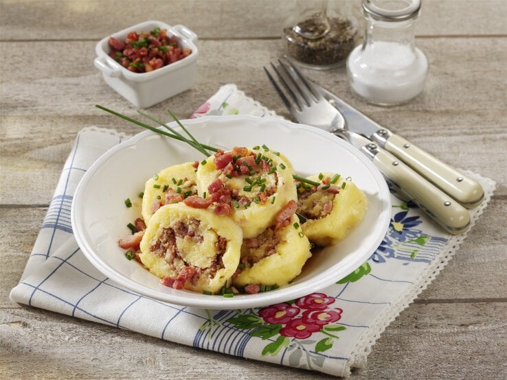 Geschnittene sächsische Wickelknödel auf einem weißen Teller auf einem bedruckten Geschirrhandtuch. Die Wickelknödel sind angerichtet mit Speck und Schnittlauch. Neben dem Teller liegen Messer und Gabel. Im Hintergrund liegt eine Schale mit Speck und zwei Streuer mit Pfeffer und Salz.