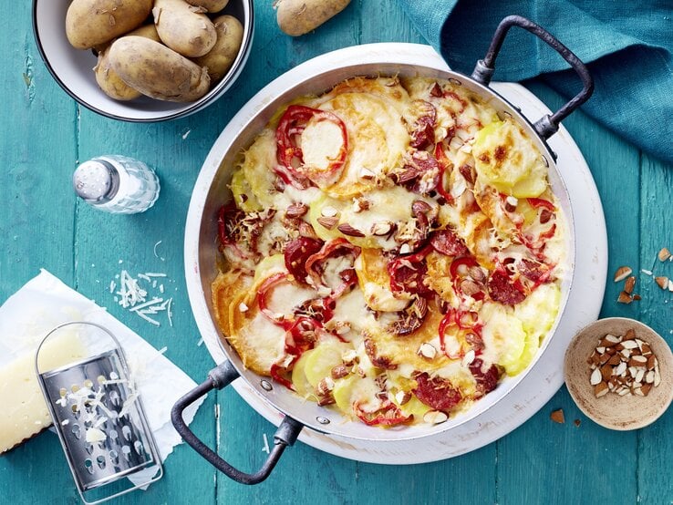 Ein spanischer Kartoffelauflauf mit Chorizo und Paprika steht auf einem blauem Untergrund. Um die Auflaufform herum liegen Käse, Kartoffeln, Salz und Mandelsplitter.