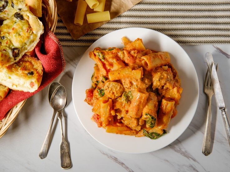 Tortelloni mit Salsiccia in Tomaten-Sahne-Soße auf weißem Teller. Daneben liegen Besteck und selbst gemachtes Knoblauchbrot. 