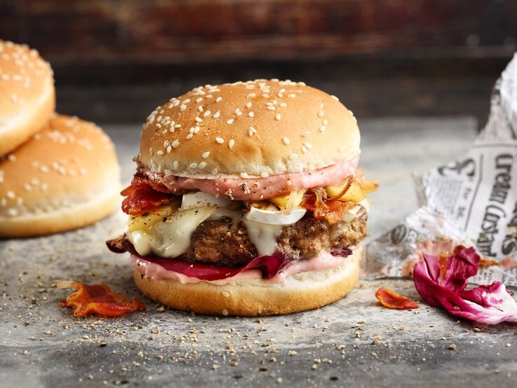 French Burger mit Speck auf einer grauen Steinplatte vor einem dunklen Hintergrund neben Zeitungspapier. 