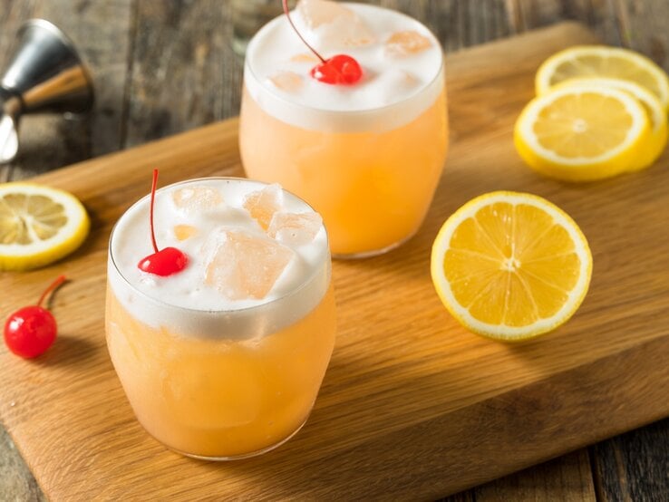 Zwei Raki Sour mit Cocktailkirsche auf Holzbrett auf Holztisch. Zitronenscheiben als Dekoration auf dem Brett verteilt. Liegendes Barmaß am oberen Bildrand. Aufnahme aus der Draufsicht.