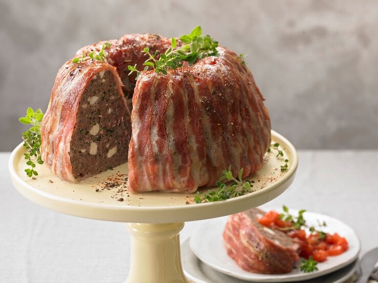 Der deftige Gugelhupf mit Rinderhack und Feta im Speckmantel garniert mit Kräutern angerichtet auf einem Kuchenständer vor einem grauen Hintergrund. 