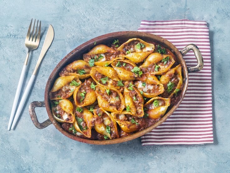 Muschelnudel-Pfanne mit Hackfleisch in einer Auflaufform. Daneben ein Küchentuch und Besteck. Das Ganze auf einem blauen Untergrund. 