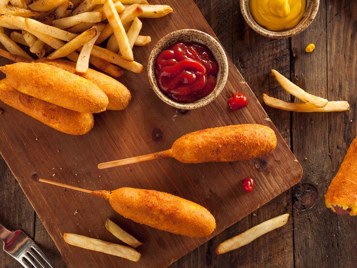 Corn-Dogs mit Pommes auf einem Holzbrett. Daneben kleine Schüssel mit Ketchup und Senf. 