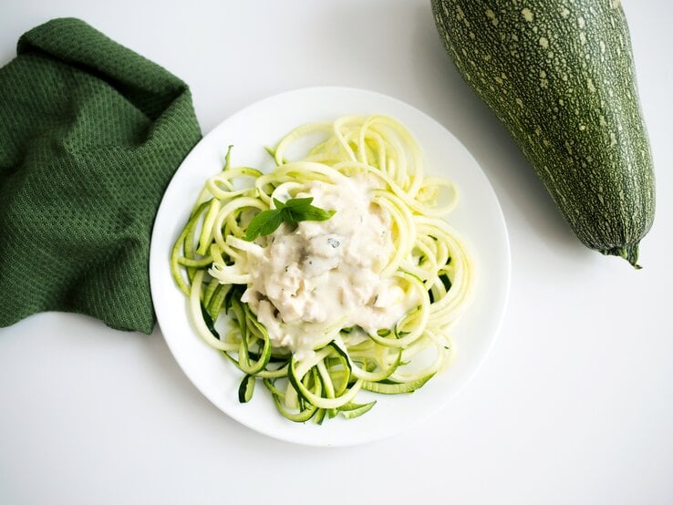 Auf einem weißen Untergrund liegen ein grünes Tuch und eine Zucchini. Daneben steht ein weißer Teller mit Zucchini-Nudeln mit Ziegenkäse-Soße. | © Shutterstock / Megan Betteridge