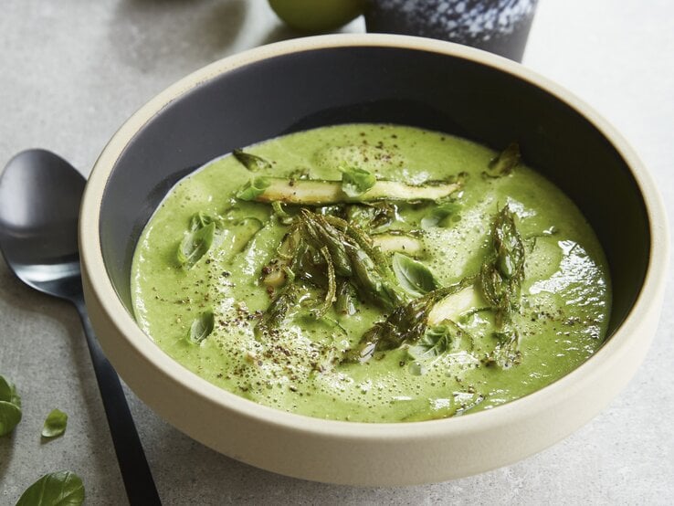 Eine graue Schale auf einem grauen Tisch mit der grünen Spargelcemesuppe. Alles von schräg oben fotografiert.