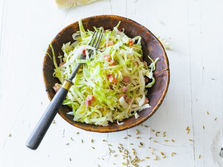 Draufsicht auf einen Bayrischen Krautsalat mit Speck in Schale. Das Ganze steht auf einem hellen Tisch. 