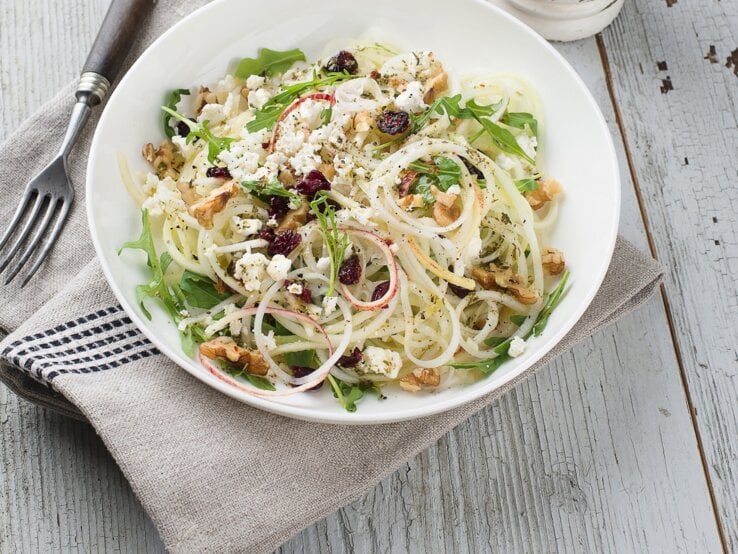 Draufsicht: Ein runder Teller mit einem Kohlrabi-Apfel-Salat mit Käse und Walnüssen auf einem hellen Untergrund aus Holz.