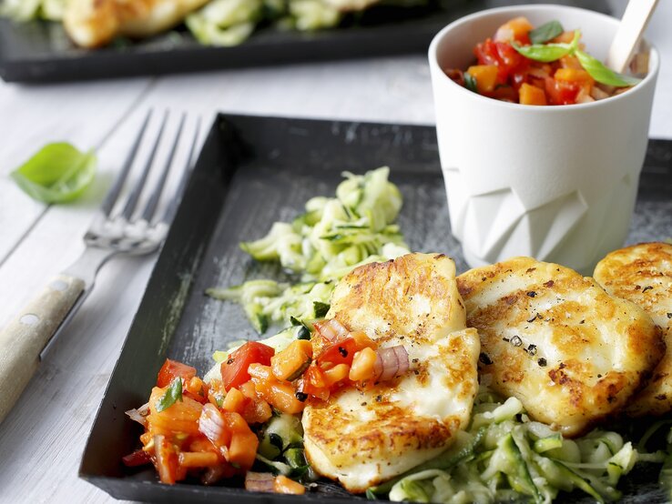 Draufsicht, schräg: Auf einem Untergrund aus hellem Holz liegt ein dunkles Tablett aus Holz. Darauf ist gebratener Halloumi auf Zucchinisalat.